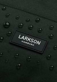 Vattentålig svart textil med droppar, med en rektangulär, präglad svart etikett som står "LARKSON HAMBURG". Strukturerad yta.
