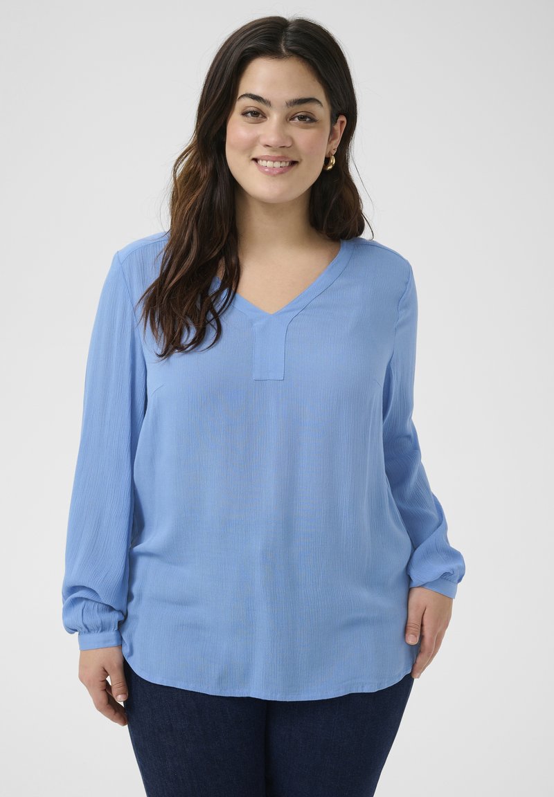 Blouse à manches longues bleu clair avec un col en V et une texture subtile. Présente une coupe décontractée et des poignets froncés. Associée à un jean foncé.