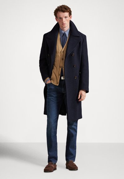 Mężczyzna w granatowym dwurzędowym płaszczu, beżowym swetrze warkoczowym, niebieskiej koszuli z krawatem, ciemnych dżinsach i brązowych zamszowych mokasynach stoi na białym tle.