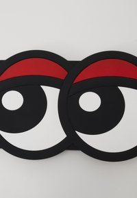 Tapis en caoutchouc noir avec deux grands yeux dotés d'iris blancs et de pupilles noires, surmontés d'accentuations semi-circulaires rouges.