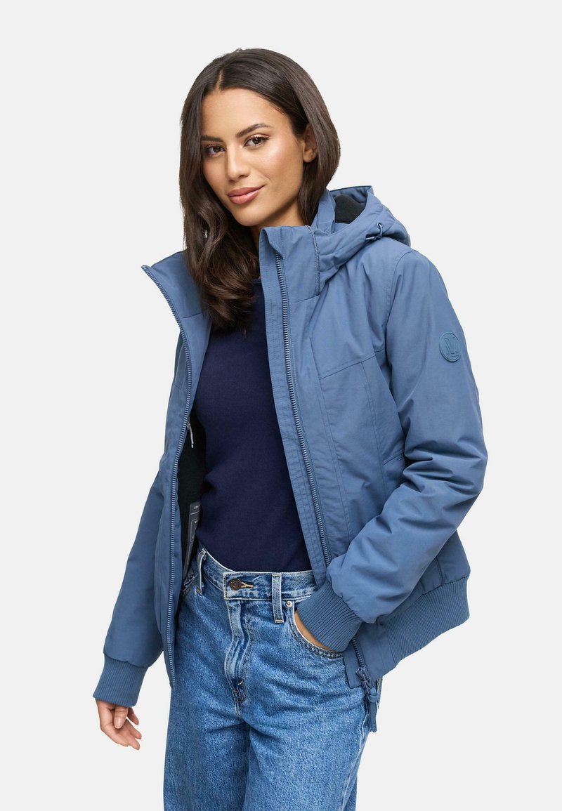 Veste légère bleue avec capuche, zip frontal, poignets côtelés et un patch rond sur la manche, portée sur un haut sombre et un jean.