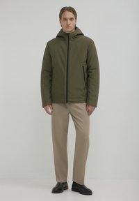 Olivgrüne gepolsterte Jacke mit Reißverschluss, Kapuze und seitlichen Taschen, kombiniert mit beigen Hosen und schwarzen Schuhen. Glatte Textur, minimalistisches Design.
