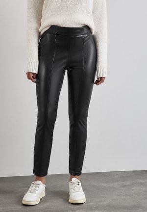 Zwarte faux leren broek met een hoge taille, voorzien van voornaad en een slim fit, gecombineerd met witte sneakers en een lichte trui.