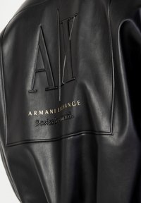Svart läderjacka med präglad "AX" logotyp, guld "Armani Exchange" text och "Basics by amani" detalj på baksidan. Slät textur.