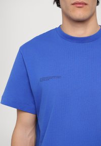 Blue organic cotton t-shirt featuring a crew neck, short sleeves, and a printed text detail on the left chest area. Smooth texture.
