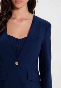 Žena v tmavě modrém blejzru se strukturovanými rameny a zlatým knoflíkem, přes odpovídající tmavě modré top s zipem.