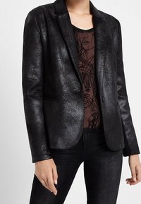 Blazer noir en simili cuir avec une texture lisse, fermeture à un bouton et revers crantés, porté sur un top à motifs.