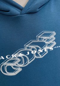Zils kapucis džemperis, kas izgatavots no mīksta kokvilnas maisījuma, ar izteiksmīgu balto grafiku "CORE" un "JACK & JONES 1990", kas izprintēta pa priekšu.