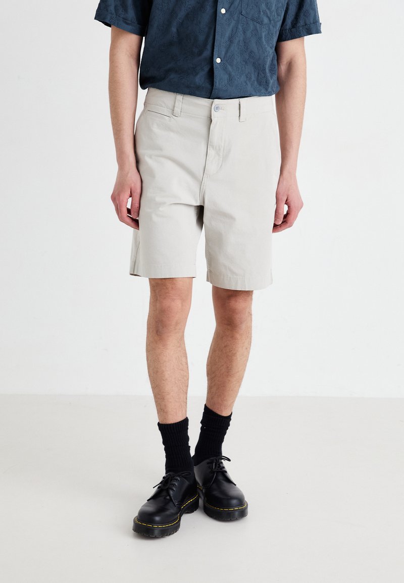 Bermudas de algodón de color gris claro con corte recto, que presentan bolsillos laterales y cierre de botón, combinadas con calcetines y zapatos negros.