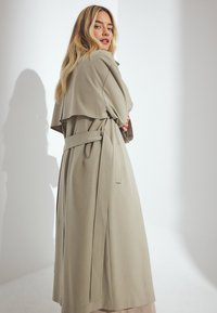 Garšs, bēšs mētelis ar dubultās rindas dizainu, platām atlokiem un jostotu vidukli. Iespējamas sānu kabatas un plūstoša silueta.