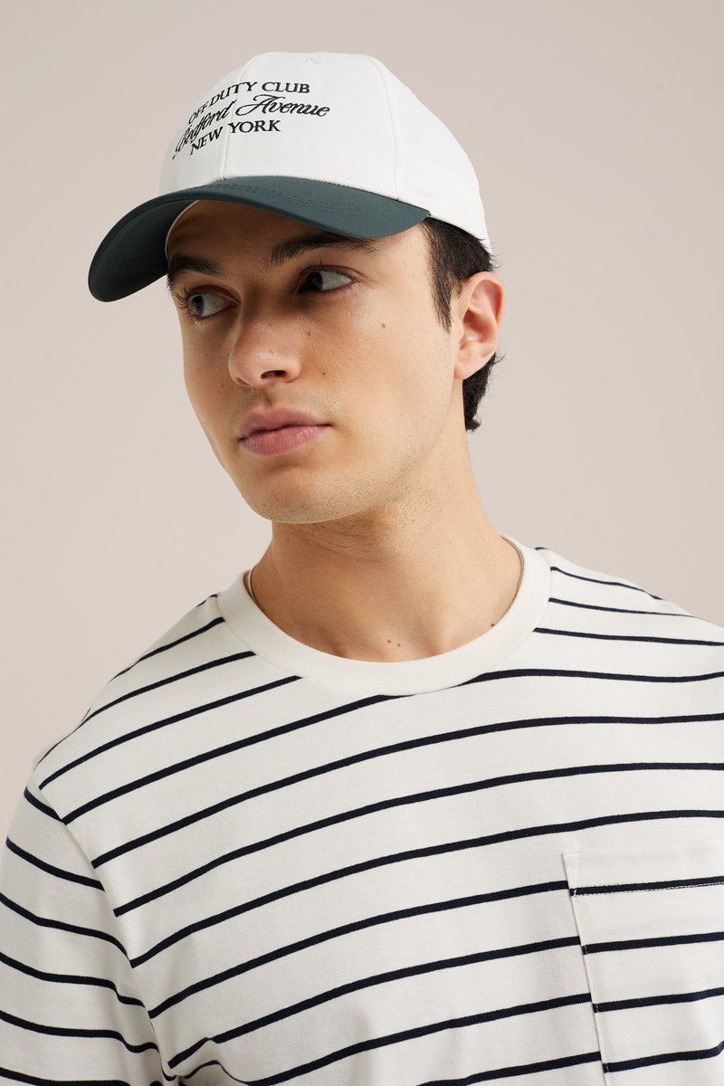 Jeune homme portant un t-shirt rayé blanc et noir et une casquette de baseball blanche avec le texte « Off Duty Club Westford Avenue New York ».
