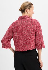 Kurze Jacke aus strukturiertem rosa Stoff mit Kragen, Dreiviertelärmeln und einem Knopfdetail an den Manschetten. Schwarze Hose sichtbar.