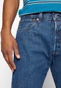 Vaqueros azules de mezclilla con corte recto, diseño de cinco bolsillos, costuras visibles y bragueta de botones. Mano apoyada en la cintura.