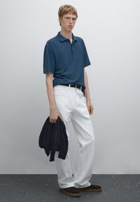 Tmavě modré polo tričko, bílé rovné džíny a hnědé nazouvací boty. Drží tmavý kardigán; jednoduchý, čistý design bez vzorů.