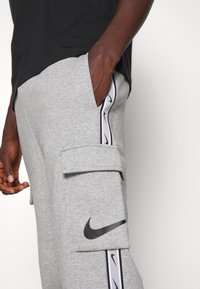Grå sweatpants i bomull med sidfickor, en cargoficka och en svart Nike-logotyp med ett vitt randmönster längs sidorna.