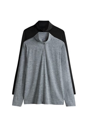 Mladý muž si upravuje čierny športový top s dlhým rukávom a predným zipsom, stojaci pred jednofarebným bielym pozadím.