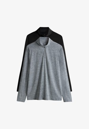 Mladý muž si upravuje čierny športový top s dlhým rukávom a predným zipsom, stojaci pred jednofarebným bielym pozadím.