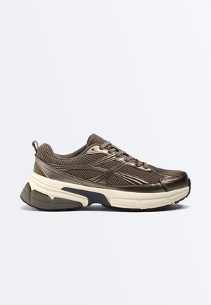 Hnedá sieťovaná a syntetická nízka teniska s hrubou bielou a čiernou štruktúrovanou podrážkou, šnurovací predok a slučka na päte na jednoduché obúvanie.