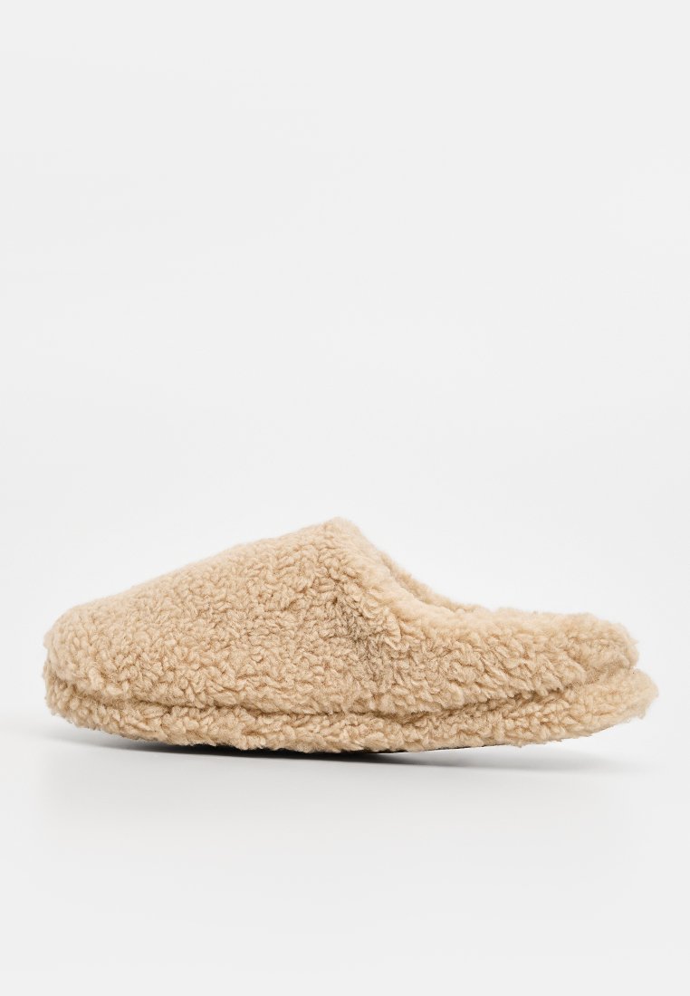 Chausson beige et duveteux fabriqué en tissu doux et texturé. Présente un design à bout fermé et à dos ouvert avec une semelle plate.