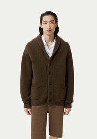 Brun stickad cardigan med sjalkrage, svarta knappar och framfickor; bärs över en vit skjorta och kombineras med bruna shorts.