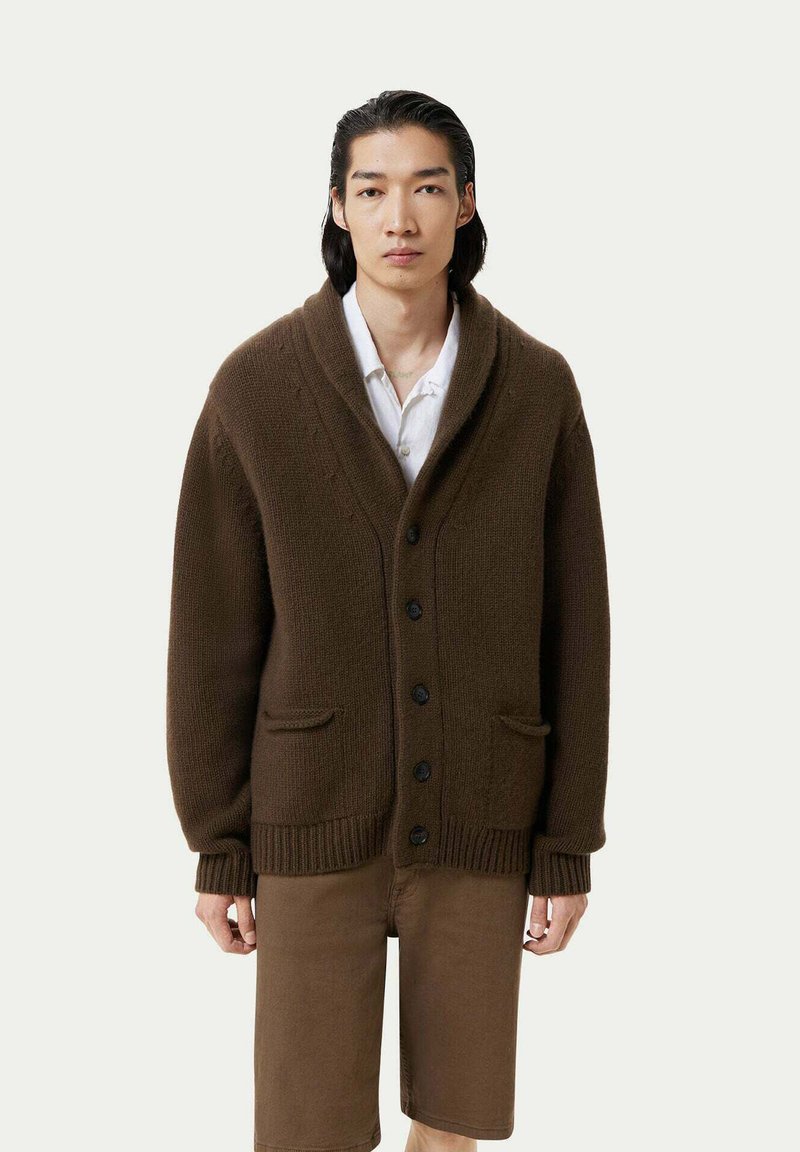 Brun stickad cardigan med sjalkrage, svarta knappar och framfickor; bärs över en vit skjorta och kombineras med bruna shorts.