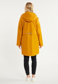 Persona con un impermeable con capucha amarillo mostaza, pantalones negros y zapatillas blancas, de pie de espaldas sobre un fondo blanco liso.