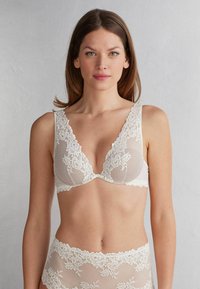 Vit spets-bandeau med djup V-ringning, blommönster, vågiga kanter och genomskinliga mesh-partier. Bandeau matchas med passande spets-trosor.