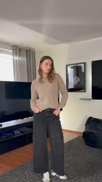 Suéter bege, jeans cargo pretos de perna larga, sapatilhas brancas com riscas pretas. Brincos grandes em argola dourada. Ambiente interior com decoração moderna.