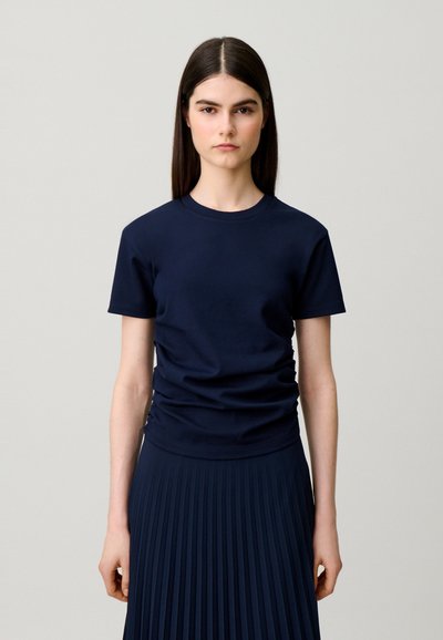 Giovane donna con lunghi capelli scuri che indossa una maglietta blu navy a maniche corte tinta unita e una gonna plissettata abbinata su uno sfondo chiaro.