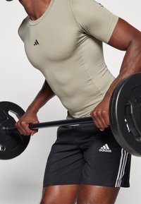 Dopasowana oliwkowa koszula z krótkim rękawem z czarnym logo, połączona z czarnymi szortami z białymi paskami i logo, podczas podnoszenia sztangi.