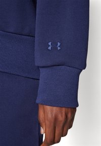 Marinblå sweatshirt med ribbade manschetter. Har en broderad mörkblå logotyp på ärmen. Tyget verkar slätt med en hållbar textur.