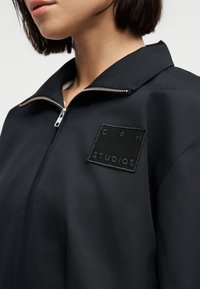 Γυναίκα που φοράει μαύρο μπουφάν με φερμουάρ και ένα μικρό τετράγωνο σημάδι με την επιγραφή "CPH STUDIOS" στο στήθος, γιακάς ελαφρώς ανοιχτός.