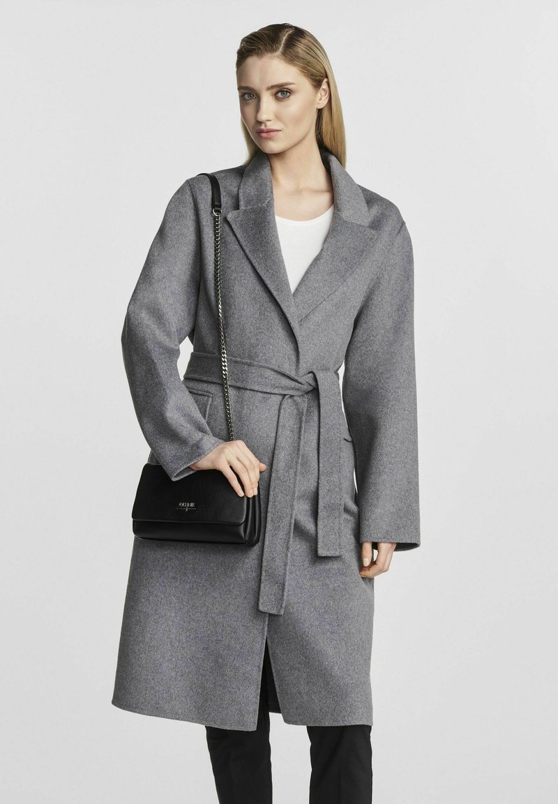 Szary wełniany płaszcz z szerokimi klapami, z paskiem w talii i długimi rękawami. W połączeniu z czarną torebką o teksturowanej powierzchni i białą bluzką. Prosty fason.