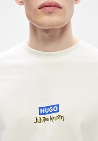 Κρεμ μπλούζα από βαμβάκι με μπλε λογότυπο "HUGO" και χρυσό κείμενο "Jujutsu Kaisen". Στρογγυλή λαιμόκοψη και κοντά μανίκια.