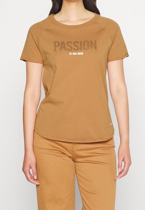 Kobieta ubrana w brązowy t-shirt z krótkim rękawem oraz dopasowane spodnie, z napisem "PASSION BY MUS MUSH" na piersi, wykonanym z kropek.