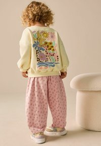 Sárga pulóver színes állat- és virágmintákkal a háton, rózsaszín pöttyös bő nadrággal párosítva. Fehér és pasztell színű sportcipők.