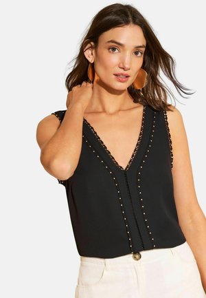 Pruunide juustega naine, kes kannab suuri vaskseid kõrvarõngaid, varrukateta musta topi kuldsete naastudega ja valgeid pükse, puudutades ühe käega oma kaela.