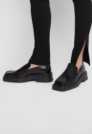 Zwarte leren instapschoenen met een chunky zool, met een gladde afwerking en minimalistisch ontwerp. De broek heeft zijsplitten voor extra detail.