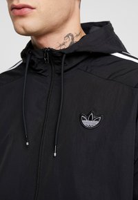 Svart lättviktsjacka med huva, med dragkedja fram, broderad logotyp på vänster bröst och vita randaccenter på axlarna.