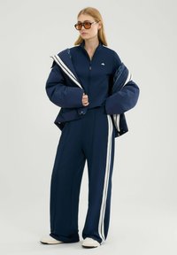 Granatowy zestaw sportowy składający się z kurtki zapinanej na zamek z białymi paskami oraz luźnych spodni. Materiał wydaje się gładki o miękkiej fakturze, w połączeniu z jasnymi butami.