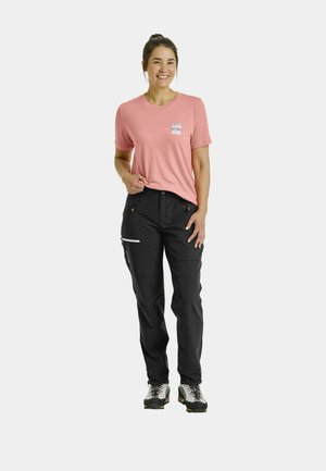 Light pink short-sleeve t-shirt with a chest logo, paired with black pants featuring zippered pockets and gray accents, worn with hiking shoes.