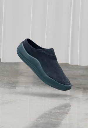Chaussure noire sans lacets avec une semelle épaisse flottant au-dessus d'un sol en béton poli, fond de rideau blanc, sans lacets ni sangles visibles.