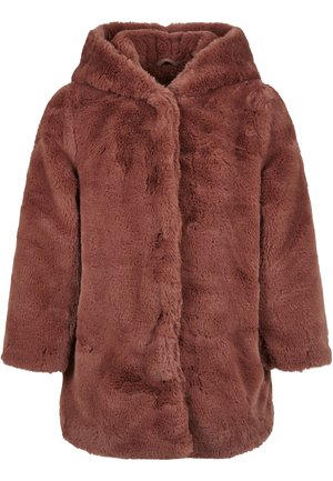 Femme portant une veste à capuche en fausse fourrure rose poudré sur un pantalon noir, avec des cheveux blonds lâchés partiellement visibles.