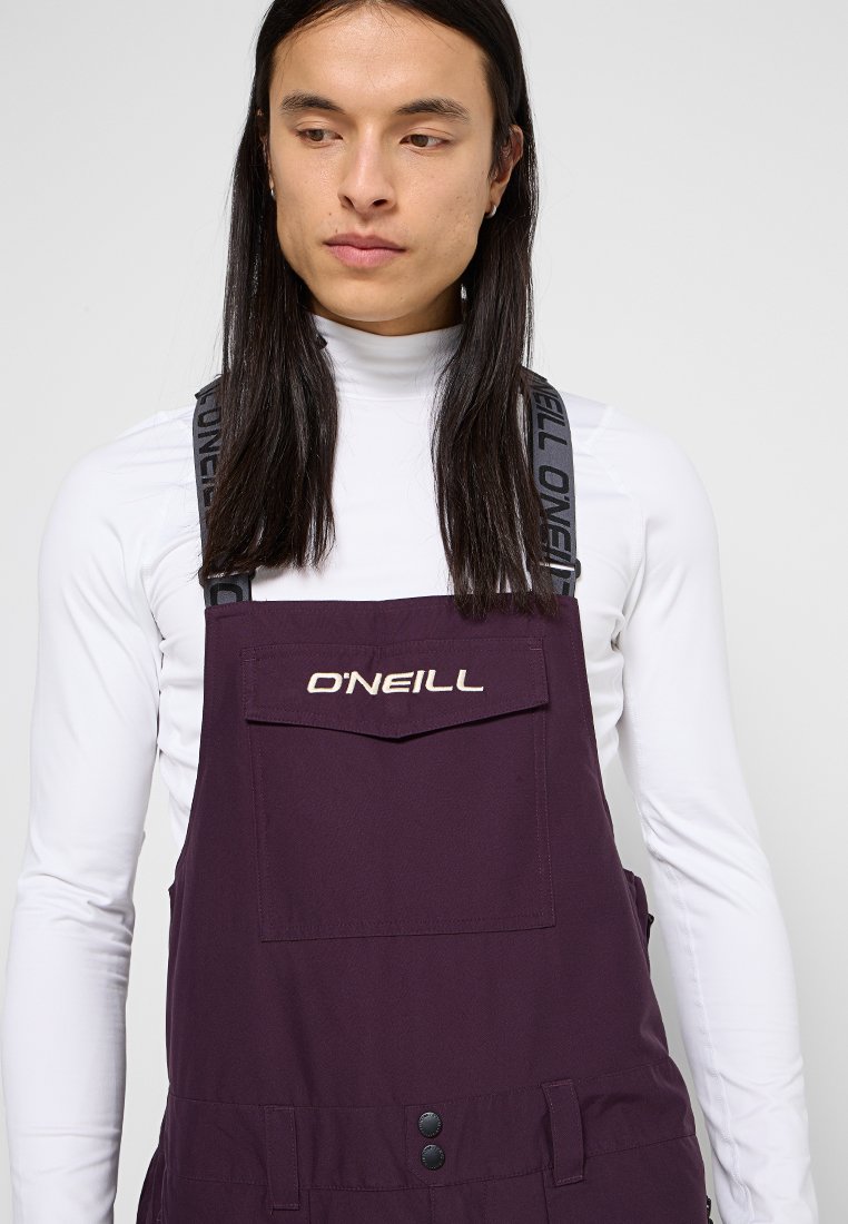 Osoba z długimi ciemnymi włosami, ubrana w białą golfową bluzkę i fioletowe ogrodniczki O'Neill z markowymi ramiączkami, patrząca w dół.