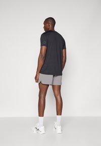 Schwarzes Kurzarmhemd mit einem Logo auf dem linken Ärmel, kombiniert mit grauen Shorts und weißen Sportschuhen, stehend vor einem schlichten Hintergrund.