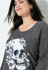 Camiseta gris de manga larga que presenta un gran gráfico de calavera rodeado de flores. Textura suave con un ajuste relajado y escote amplio.