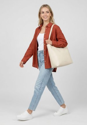 Blonde vrouw die loopt, gekleed in een roestkleurig overhemd met knopen, een wit topje, lichtblauwe spijkerbroek, witte sneakers en een grote beige tas over haar schouder dragend.