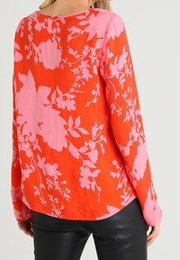 Blusa rossa con motivo floreale rosa, maniche lunghe, scollo rotondo e tessuto leggero; indossata con pantaloni neri aderenti.