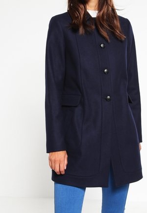 Vrouw draagt een knielange donkerblauwe jas met drie zwarte knopen, klepzakken en lichtblauwe jeans, staand tegen een witte achtergrond.