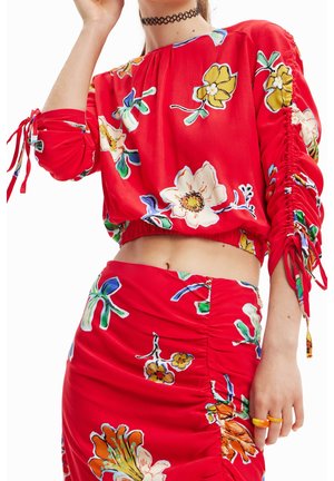 Femme portant une tenue deux pièces rouge à motifs floraux avec des côtés froncés et des liens aux manches, montrant le ventre et accessoirisée d’un collier ras-du-cou noir.
